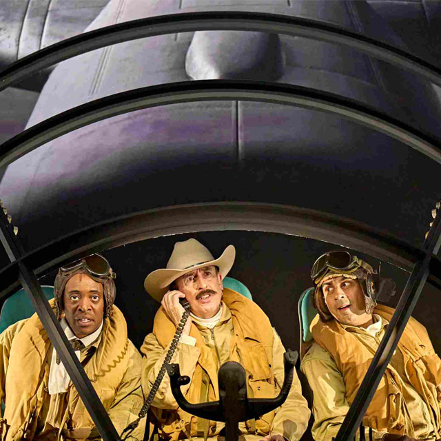 Three pilots inside a plane during a production. The middle pilot is using a wired telephone.