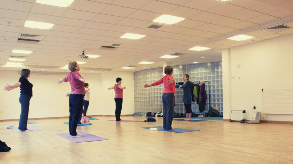 A group of people in a yoga class stood on their yoga mats alongside their yoga teacher.