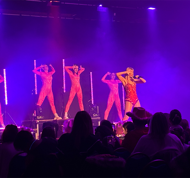 A Taylor Swift tribute act performing on stage in a red sequined outfit with back-up dancers and a big crowd watching them.